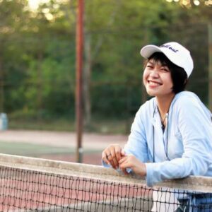 Beautiful Asian woman with short hair, wearing hat and smiling broadly on tennis court.